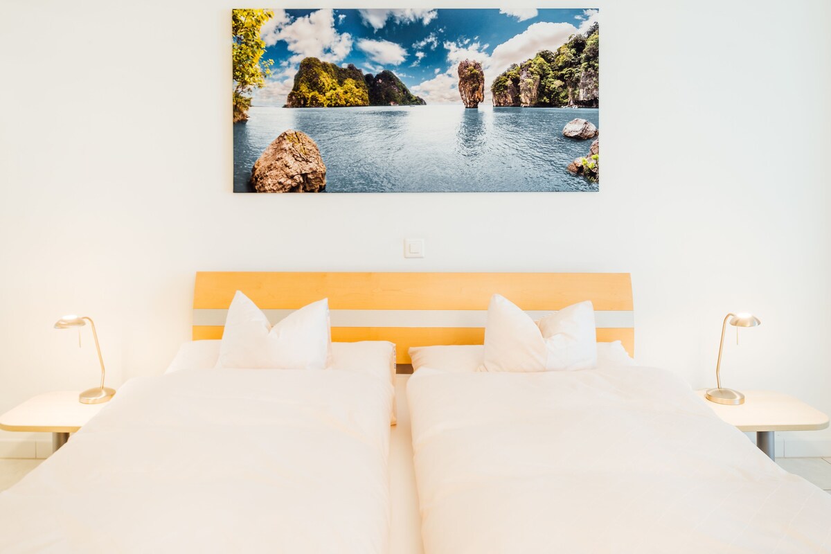 A hotel room with two beds, a large window with a view of a lake and mountains, and bedside lamps.