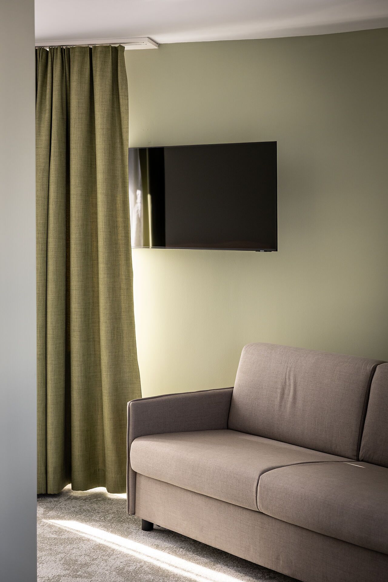 A modern living room with a sofa, a flat-screen TV mounted on the wall, and a green curtain.