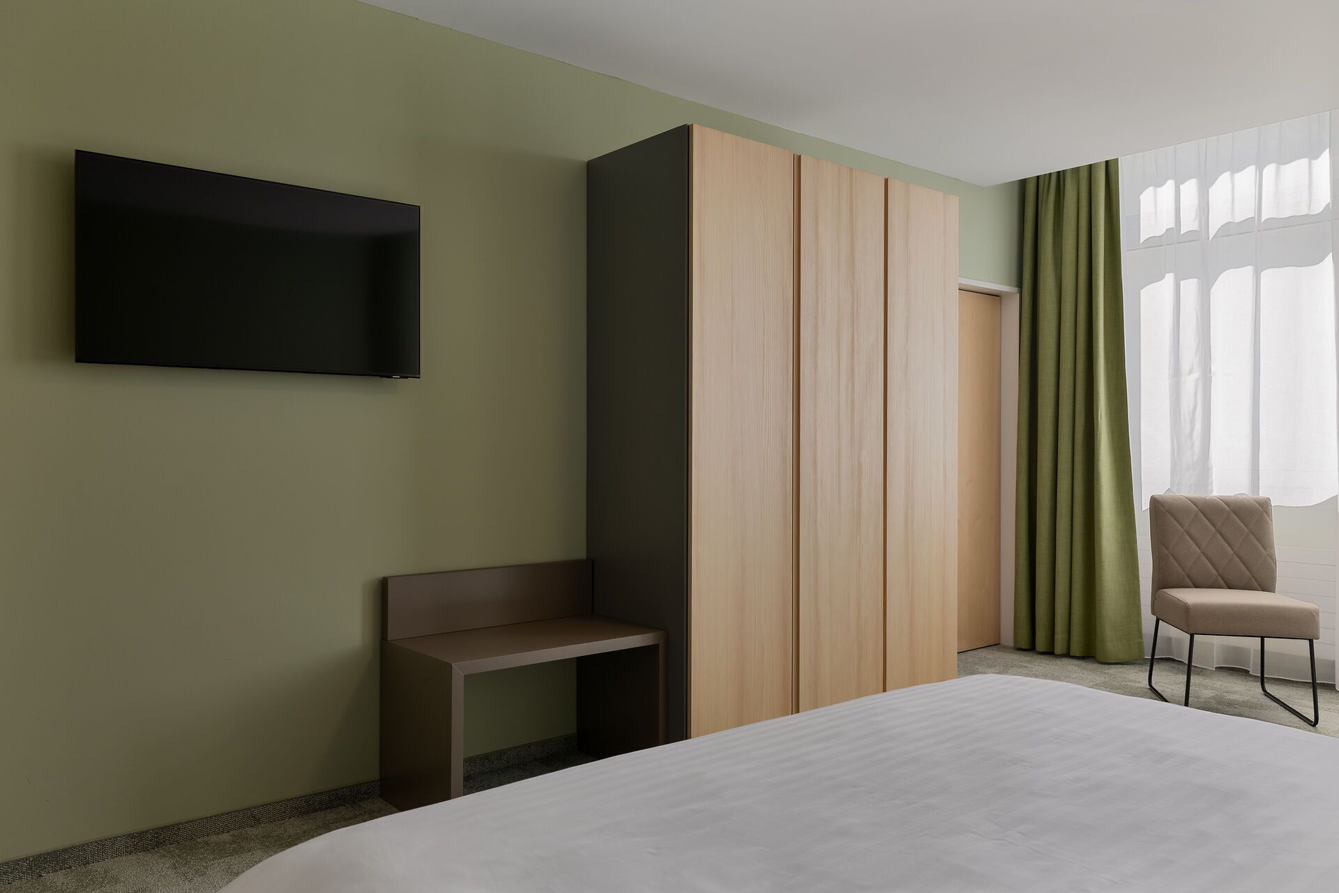 Quarto de hotel com uma cama, televisão na parede, área de estar com uma poltrona e um guarda-roupa.