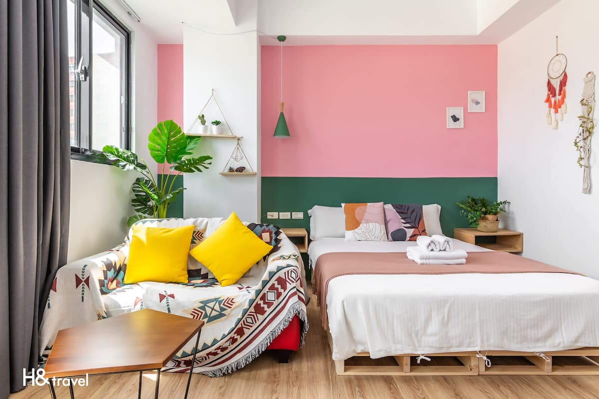 Quarto de hotel moderno com uma grande janela, uma cama com colcha branca, um sofá com almofadas coloridas e uma pequena mesa de madeira.