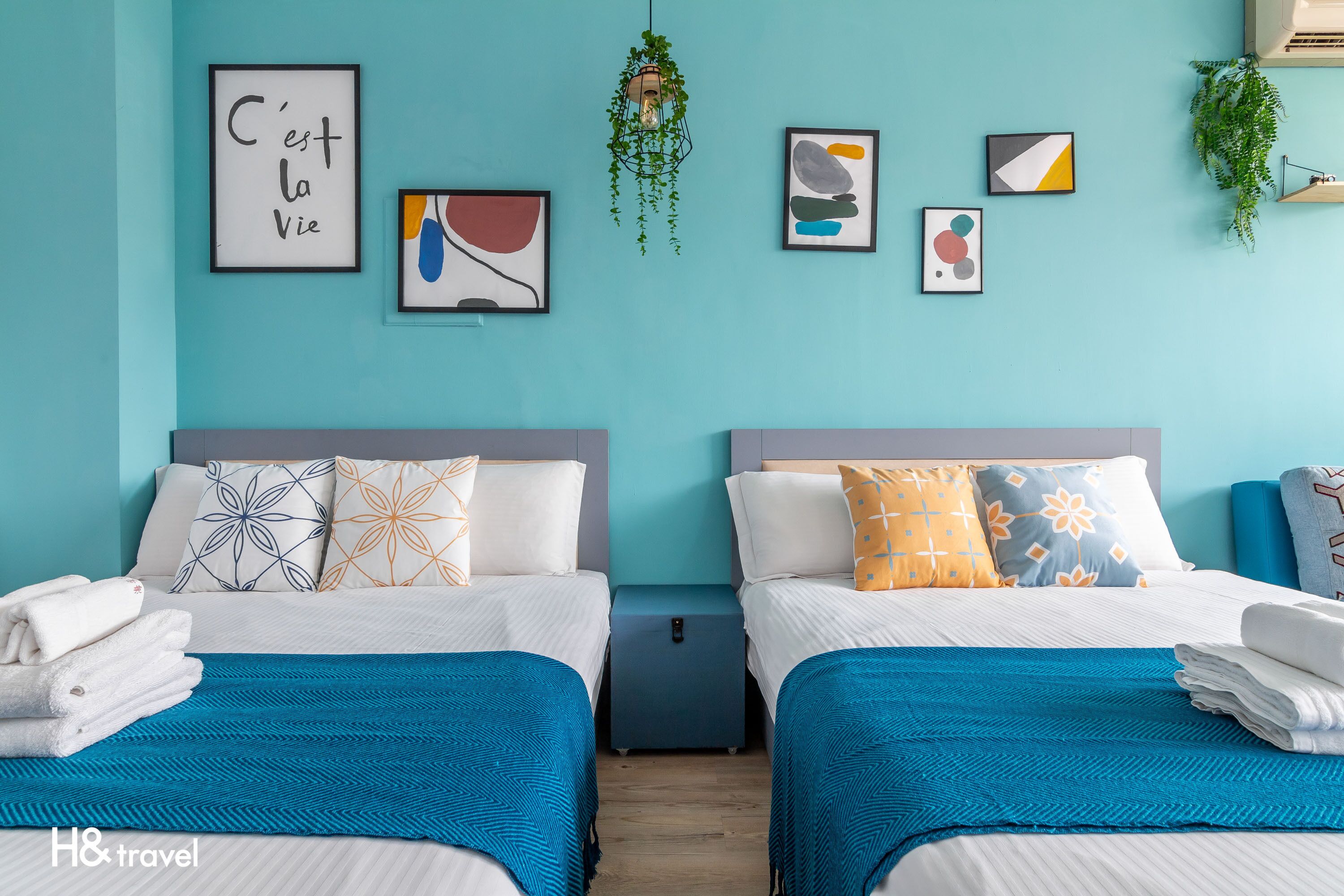 A twin room with two beds, each with a blue bedspread and white bedding. There are multiple framed artworks on the wall, a hanging plant, and a small table between the beds.