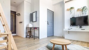 A modern, well-lit room with a wooden floor, a round wooden table, a grey cushioned bench, a small stool, a TV mounted on the wall, and a door with a grey frame. - Sleepier Multi-Minsu Tainan (Tainan)