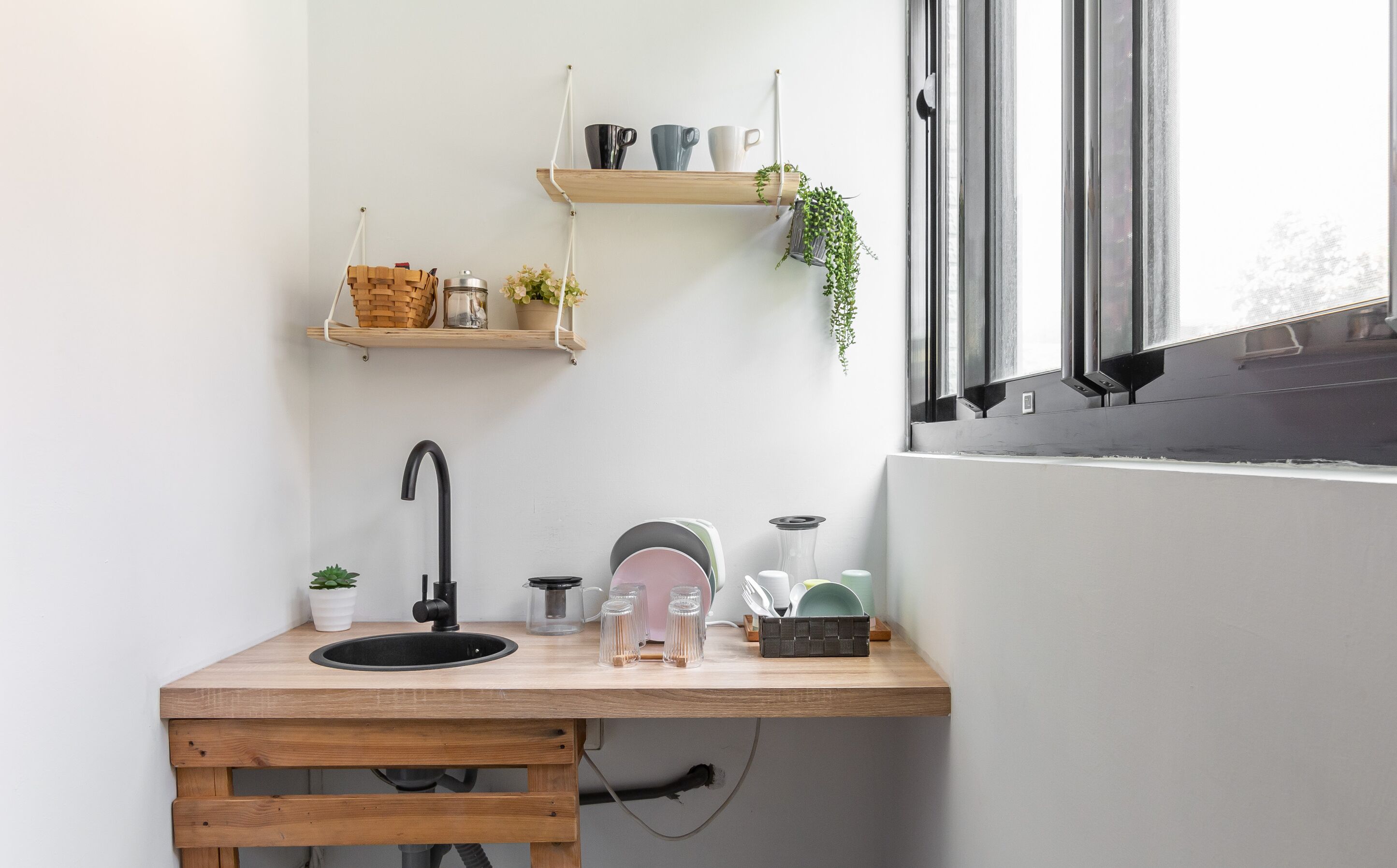 Una pequeña cocina con encimera de madera, un lavamanos y una ventana con persianas. Hay estantes sobre la encimera que albergan utensilios de cocina y una planta.