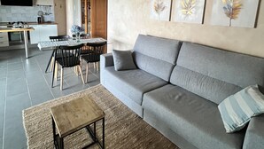 Un soggiorno moderno con un divano grigio, un tavolino da caffè e un'area pranzo con tavolo e sedie. Alle pareti si notano dipinti delicati e il soffitto è in legno.