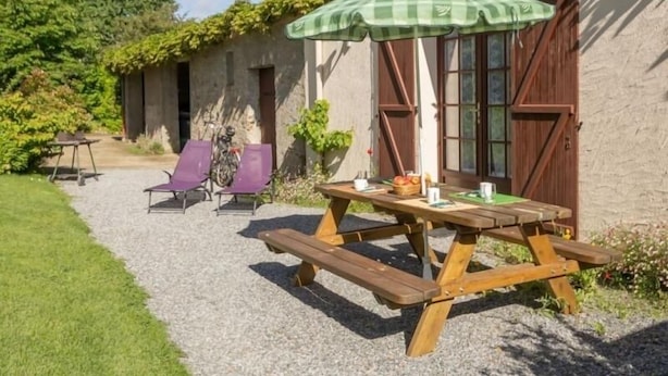 A rustic outdoor seating area with a wooden table and benches, a green checkered umbrella, and a gravel path leading to a building with a tiled roof.