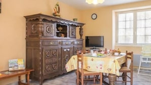 A dining area with a wooden cabinet, a table with a floral tablecloth, chairs, a bench, and a television on the wall.