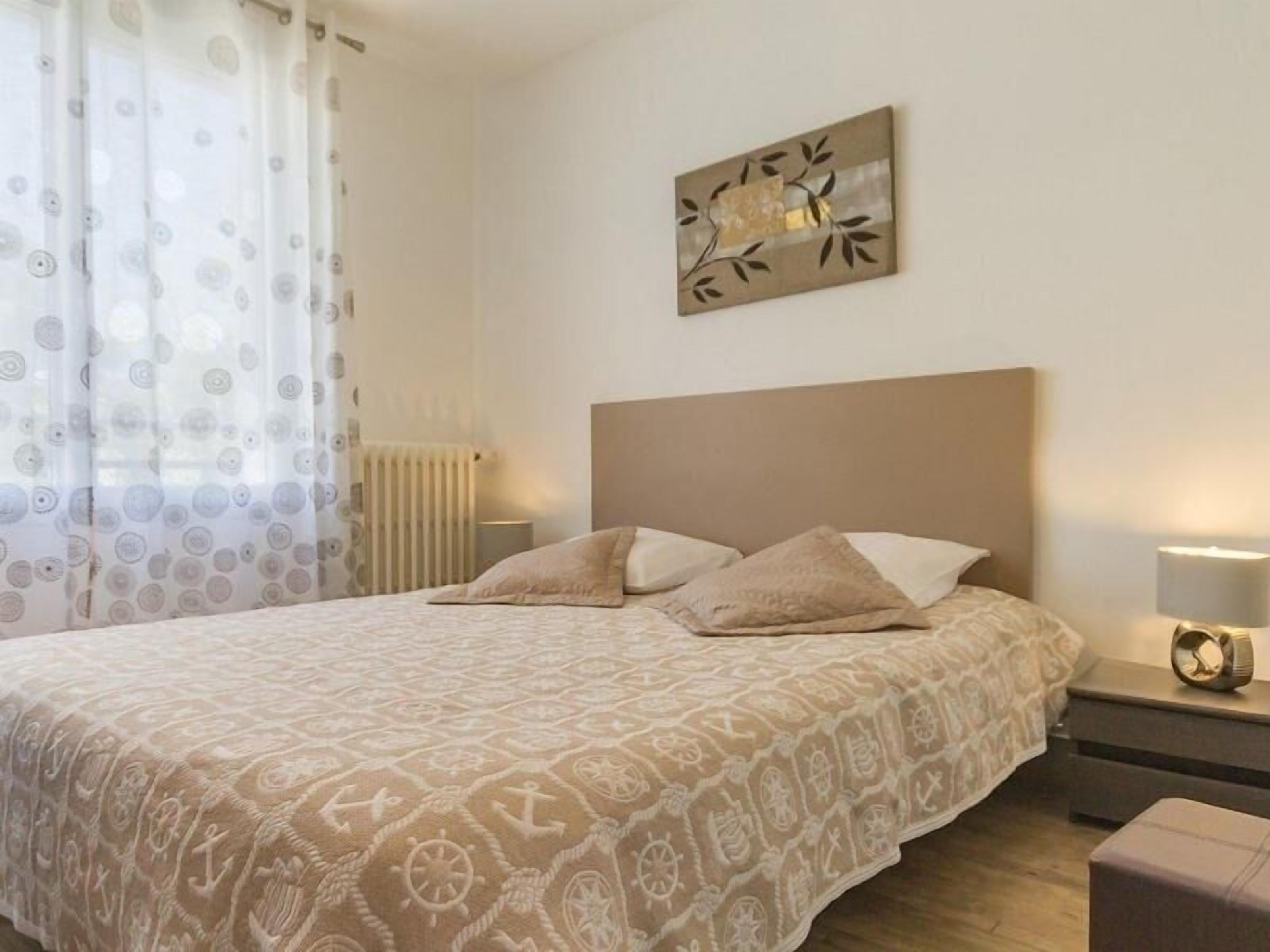 Ein ordentlich bezogenes Bett mit einem gemusterten Überwurf, ein Nachttisch mit Lampe und ein an der Wand hängendes Kunstwerk.