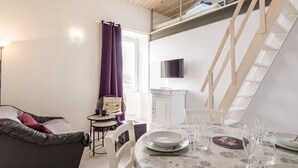 A room with a dining table set for four, a sofa, a chair, a TV on the wall, and a staircase. - Quiet gîte, WiFi, near Pornic, 4 guests (Pornic-Le Clion sur Mer)