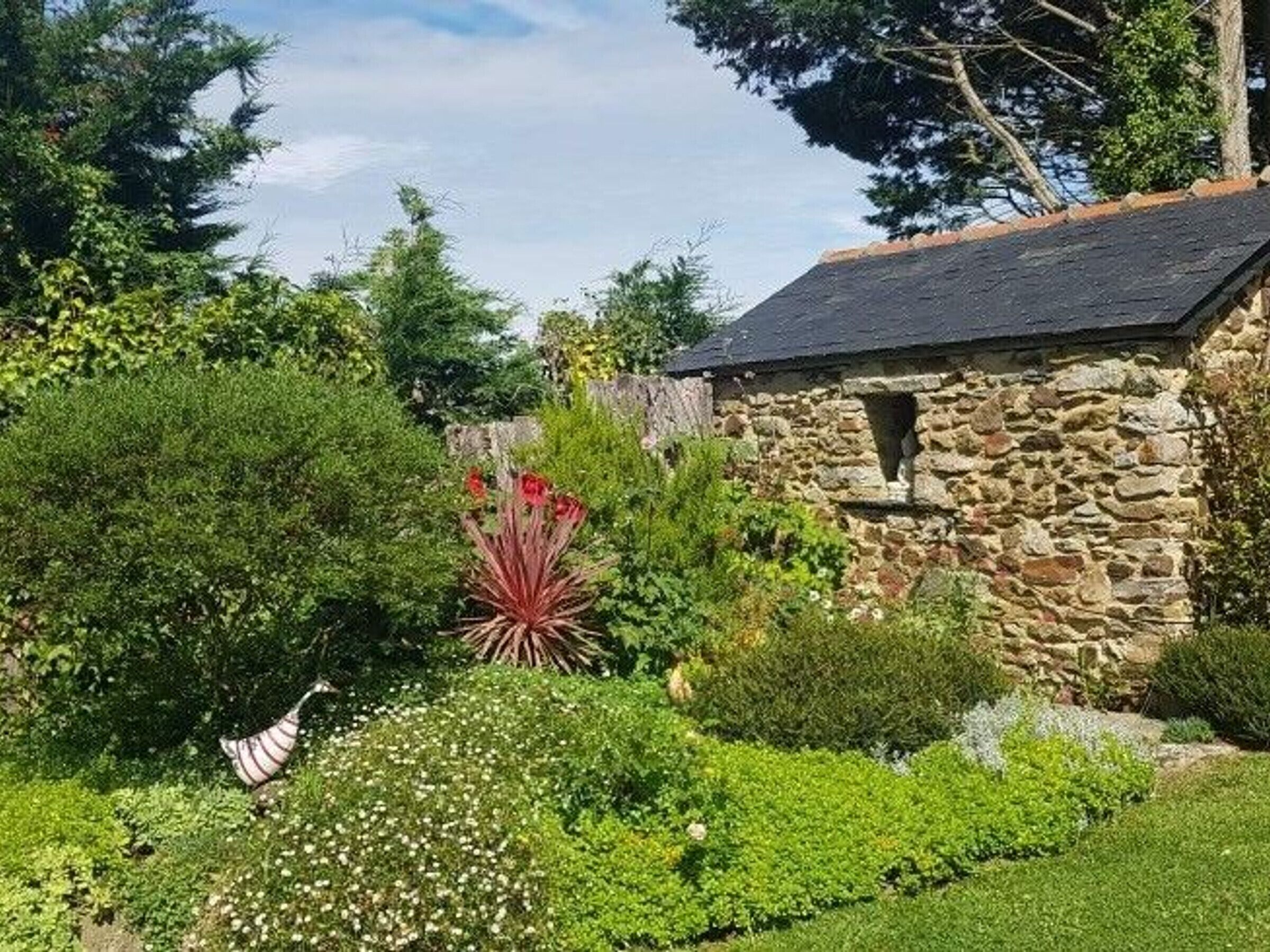 A stone building with a sloped roof, surrounded by lush greenery and a variety of plants.