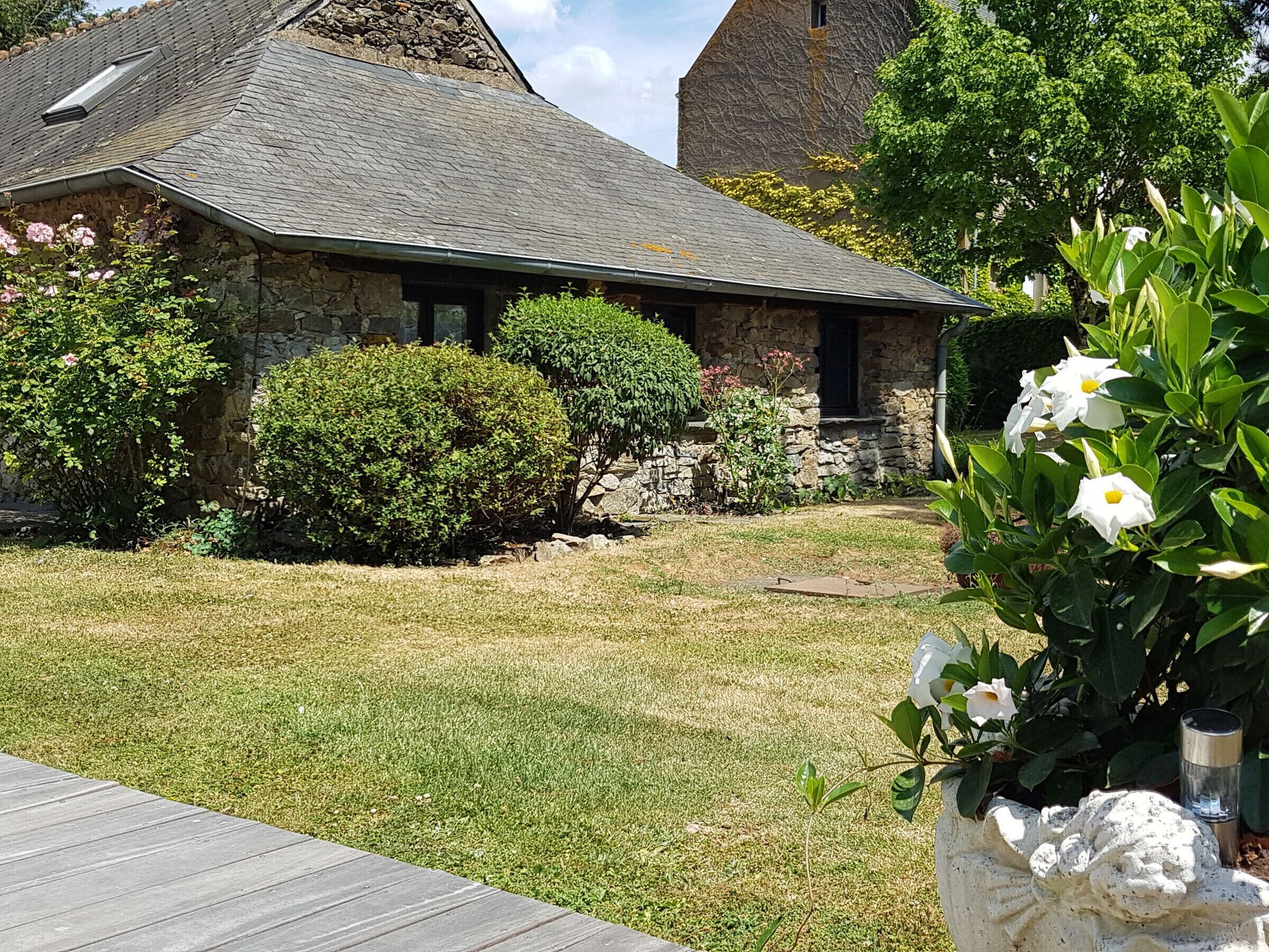 Une maison en pierre avec un toit en pente, entourée d’une pelouse bien entretenue et de divers arbustes.