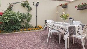 Eine Terrasse im Freien mit einem für zwei Personen gedeckten Tisch, umgeben von weißen Stühlen, einem Zierbogen und Topfpflanzen.