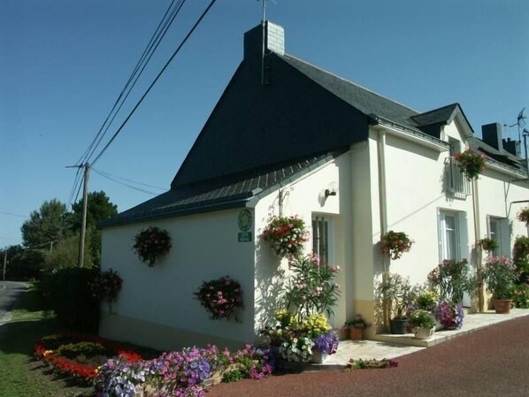 A white building with a black roof, surrounded by a variety of potted plants and flowers.