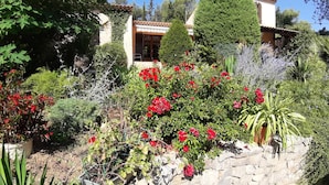 A garden with various plants and flowers, a stone wall, and a building with a tiled roof in the background. - Les chambres de Rocbaron (Rocbaron)