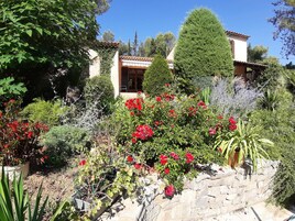 Um jardim com diversas plantas e flores, um muro de pedra e um edifício com telhado de cerâmica ao fundo.