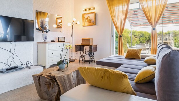 A modern living room with a flat-screen TV, a wooden coffee table, a sofa with yellow cushions, and a large window with curtains.
