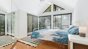 A bedroom with a sloped ceiling, large windows with blinds, a bed with a blue and white patterned bedspread, a bedside lamp, and a woven basket on the nightstand. - Paws and Relax - accommodates the whole family including your fur babies (Elizabeth Beach)
