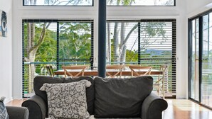 A modern living room with a grey sofa, a patterned throw pillow, and a dining area with a wooden table and chairs. Large windows with blinds allow natural light and provide a view of greenery outside. - Paws and Relax - accommodates the whole family including your fur babies (Elizabeth Beach)