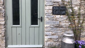 Una cabaña rústica con puerta verde, pared de piedra y un cartel que dice 'MilkChurn Cottage'.