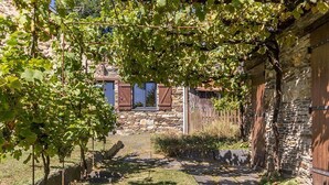 A stone building with a wooden door and window shutters, a stone wall, and a grassy area with a pathway leading to a stone building in the background. - Stone House Retreat with Garden & WiFi in Tranquil Countryside (Moisdon-la-Rivière)