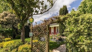 Un camí de jardí que condueix a un edifici amb una porta vermella, envoltat de vegetació exuberant i un senyal que indica una ruta ciclista.