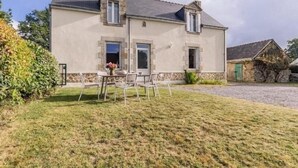 Une maison à deux étages avec une façade en pierre, un porche à l’avant avec des chaises et un petit jardin.