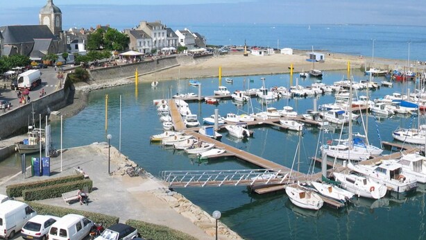 A marina with numerous boats docked, a sandy beach, and buildings in the background.