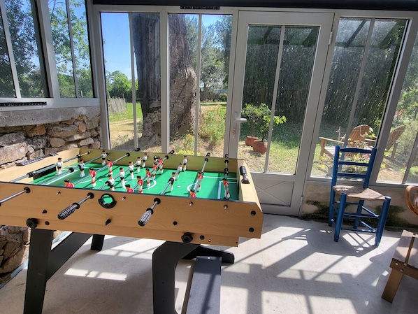 A foosball table in a sunlit room with a stone wall and a view of a garden through large windows.