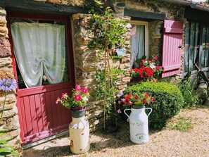 A stone cottage with a red door, a window with a curtain, and a welcome sign on a white pitcher.