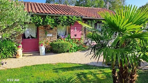 A stone house with a red-tiled roof, a red door, and a small garden with a palm tree. - Seaside Eco Cottage: 18th-Century Stone House near Pornic, 15min to Beach, Accessible, Private Garden (Arthon-en-Retz)