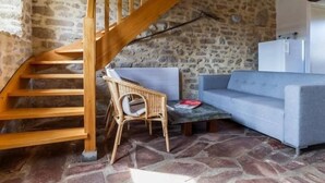 A rustic interior with a stone wall, a wooden staircase, a wicker chair, a blue sofa, and a small table. - Charming Cottage Near Salt Marshes & Guérande Walls with Enclosed Garden and Terrace (Guérande)