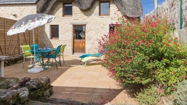A stone house with a thatched roof, a patio with outdoor furniture, and a flowering bush. - Charming Cottage Near Salt Marshes & Guérande Walls with Enclosed Garden and Terrace (Guérande)