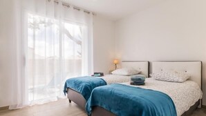 Une chambre avec deux lits, une grande fenêtre avec des rideaux légers et une table de chevet avec une lampe.