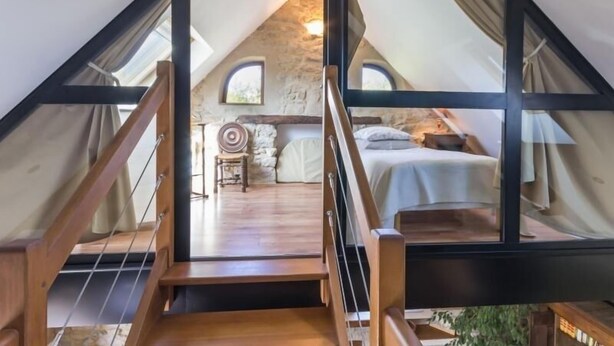 Une chambre confortable située dans les combles, avec un escalier en bois, un mur en pierre et un lit recouvert d’une couverture blanche.