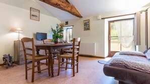 A cozy room with a wooden dining table, four chairs, a bed with patterned bedding, a radiator, and a TV on the wall. - Charming Renovated Farmhouse with Pond, Near Nantes - WiFi, Baby Equipment Included (Corcoué-sur-Logne)