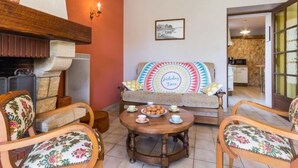 Un salon chaleureux avec une cheminée, un canapé agrémenté de coussins colorés et une table à manger mise avec des tasses à thé et des pâtisseries.