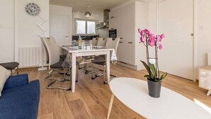 A modern living room with a dining area, a kitchen in the background, and a potted orchid on a white table. - Beachfront Apartment with Stunning Sea Views, Wi-Fi, and BBQ Area (Piriac-sur-Mer)