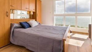 A bedroom with a bed, wooden wardrobe, and a large window with a view of the sea. - Beachfront Apartment with Stunning Sea Views, Wi-Fi, and BBQ Area (Piriac-sur-Mer)