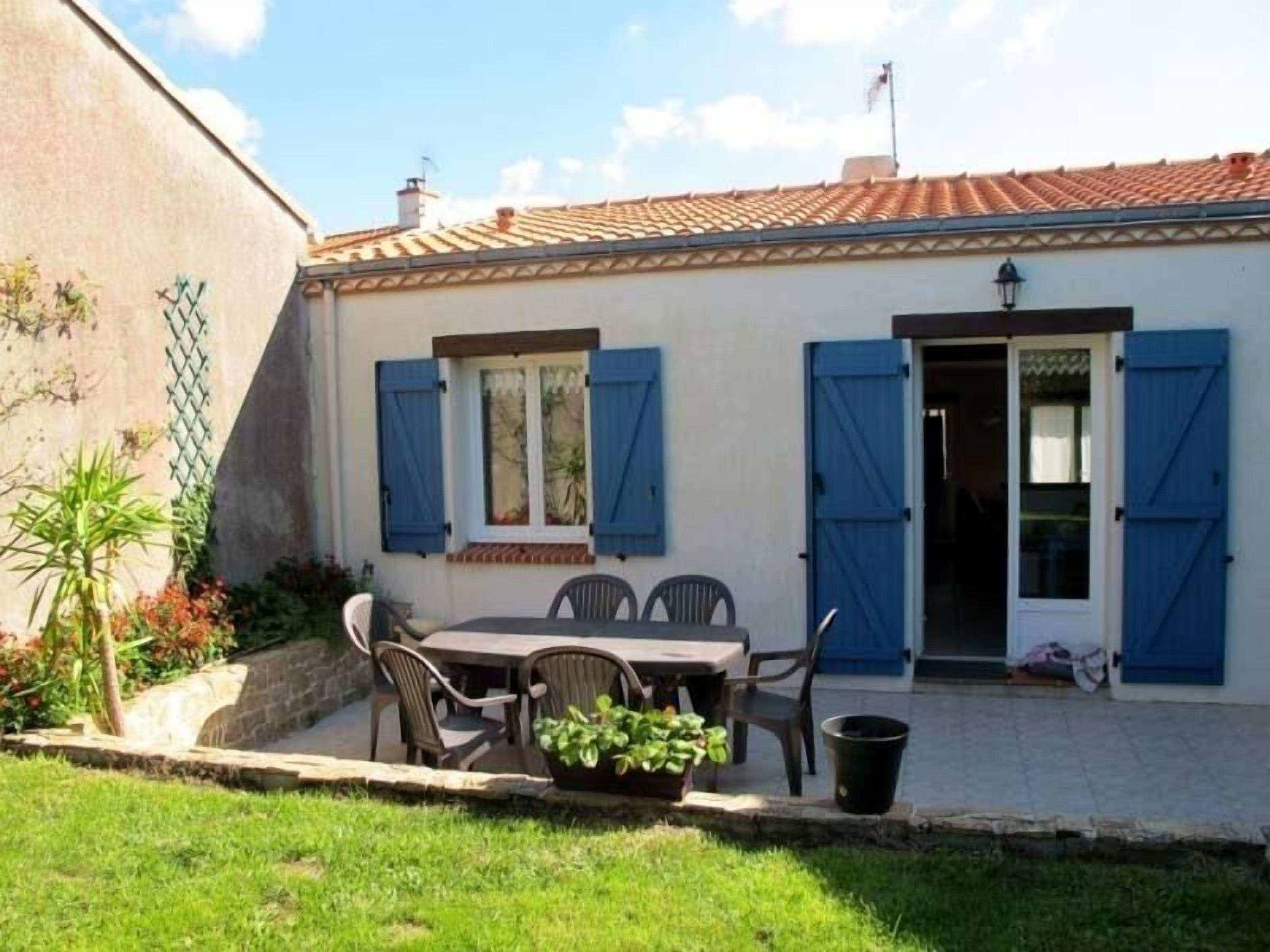 A small house with a tiled roof, a patio with a table and chairs, and a door with blue shutters.