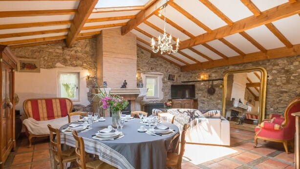Un comedor con una mesa redonda dispuesta para seis personas, rodeada de sillas de madera. Se observa una chimenea de piedra y un televisor al fondo.