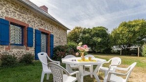 Un edificio de piedra con contraventanas azules, una mesa blanca con sillas y un jarrón de flores.