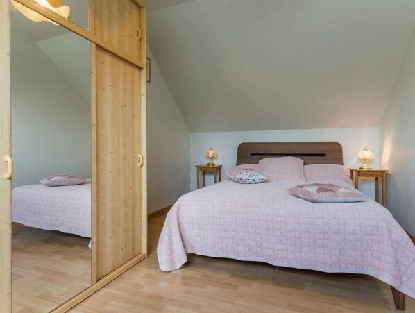 A bedroom with a wooden wardrobe, a bed with a quilted bedspread, two bedside lamps, and a mirror.