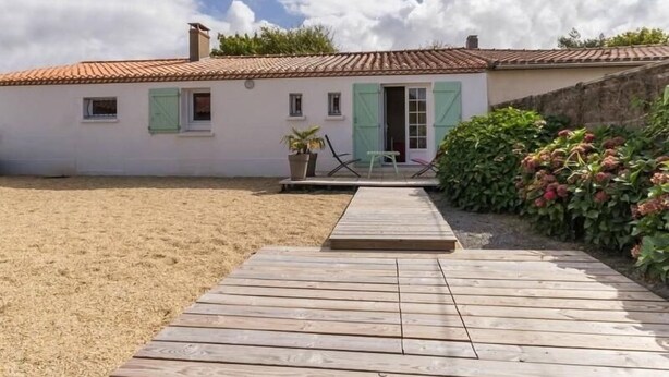 Ein weißes Gebäude mit einem Ziegeldach, einer Holzterrasse und einem kleinen Gartenbereich mit Topfpflanzen und einem Stuhl.
