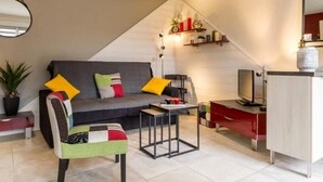 A modern living room with a grey sofa, colorful cushions, a small table, a chair, a TV, and a decorative shelf. - Charming Renovated Cottage in Medieval Clisson with Terrace, Garden, and Modern Amenities (Clisson)