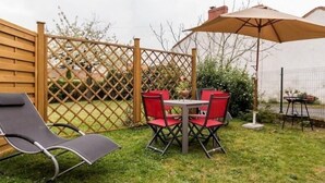 A backyard with a wooden fence, a table with four red chairs, a lounge chair, and a large umbrella. - Charming Renovated Cottage in Medieval Clisson with Terrace, Garden, and Modern Amenities (Clisson)