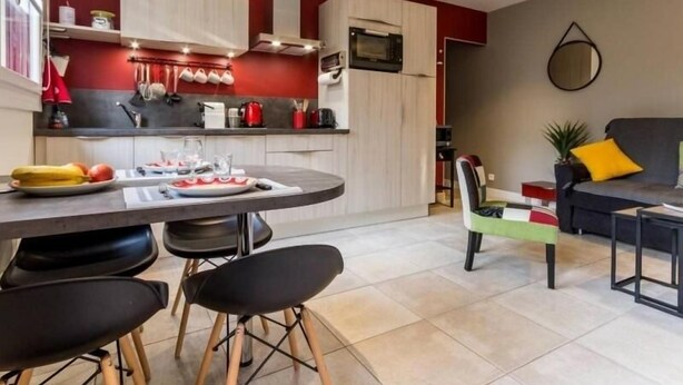 A modern kitchen with a red accent wall, a dining area with black chairs, and a grey sofa in a living room. - Charming Renovated Cottage in Medieval Clisson with Terrace, Garden, and Modern Amenities (Clisson)