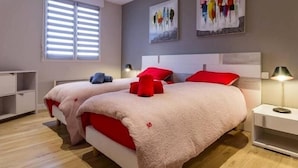 A modern bedroom with two beds, a window with blinds, artwork on the wall, and a bedside table with a lamp. - Charming Renovated Cottage in Medieval Clisson with Terrace, Garden, and Modern Amenities (Clisson)
