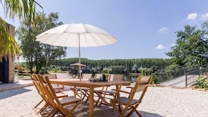 Une table ronde en bois, entourée de six chaises, et un grand parasol, installés sur une zone sablonne d’où l’on peut admirer un lac et des arbres en arrière-plan.
