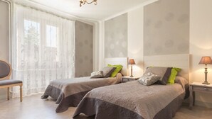 Quarto de hotel com duas camas, uma cadeira, janela com cortinas leves e um lustre.
