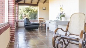 Uma área de varanda coberta com parede de tijolos, uma cadeira preta, uma cadeira de vime e uma mesa com um vaso de planta.