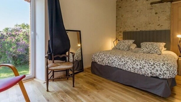 A bedroom with a large bed, a chair, a window with a view of greenery, and a stone wall. - Charming Cottage in Vineyards - Near Nantes & Clisson - WiFi, Family-Friendly (Monnières)